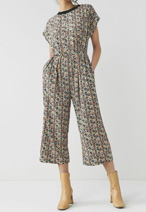 Femme portant une combinaison à motif floral avec des manches courtes et des jambes larges et raccourcies, associée à des bottines beiges.