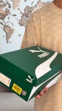 Asmuo, vilkintis smėlio spalvos tekstūruotu megztiniu, laiko žalią Puma batų dėžutę, o fone neryškiai matosi pasaulio žemėlapis.