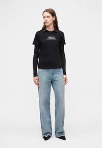 Kobieta ubrana w czarną, warstwową koszulkę polo z długim rękawem marki Armani Exchange, niebieskie szerokie spodnie dżinsowe i czarne spiczaste buty, stojąca na białym tle.