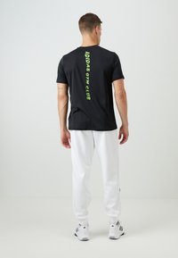 Černé sportovní tričko s nápisem "ADIDAS GYM CLUB" v jasně zeleném vertikálním tisku на zadní straně, v kombinaci s bílými atletickými kalhotami.