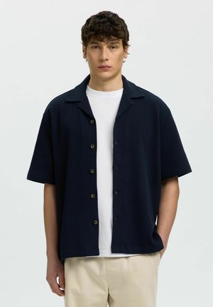 Młody mężczyzna w granatowej krótkiej koszuli na guziki, na białej koszulce i beżowych spodniach, stojący na jasnym, jednolitym tle.