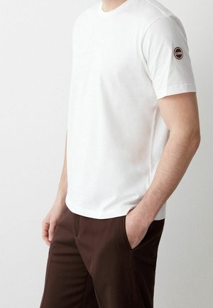 Hombre con camiseta blanca de manga corta lisa con logo redondo de Colmar en la manga y pantalones marrón oscuro, con las manos en los bolsillos, de pie frente a un fondo claro.