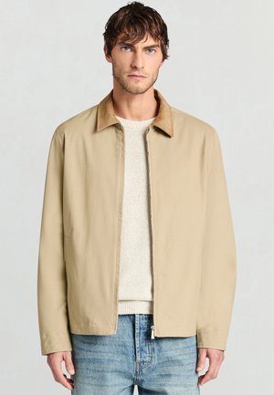 Veste beige clair avec un col en suède beige ; dotée d'une fermeture éclair à l'avant, de manches longues et d'une coupe décontractée. Portée par-dessus un pull crème et un jean bleu.