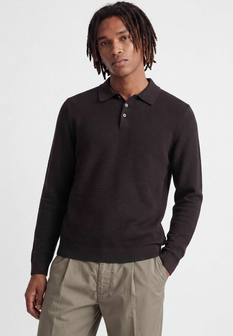 Jeune homme avec des dreadlocks mi-longs portant un polo à manches longues foncé et un pantalon beige, debout avec une main dans la poche.