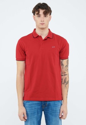 Giovane con capelli ricci che indossa una polo rossa e jeans blu, mostrando i tatuaggi sull'avambraccio destro, in piedi davanti a uno sfondo bianco semplice.