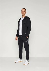 Čierna zipsová mikina s kapucňou a prednými vreckami, v kombinácii s čiernymi slim-fit nohavicami s logom, obuté s bielymi športovými topánkami.