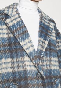 Kostkovaný kabát v modré, bílé a hnědé vlněné směsi. Má klopy s výstřihem, zapínání na jeden knoflík a měkký texturovaný povrch.