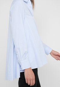 Woman wearing a light blue pinstriped long-sleeve shirt with a loose flare at the back, paired with black pants.