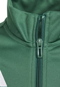 Grüne Zip-Jacke mit hohem Kragen, strukturiertem Stoff und einem Kunststoff-Reißverschluss. Verfügt über ein weißes Logo auf der linken Seite.