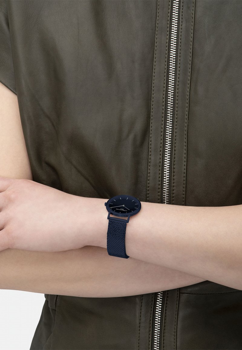 Blauwe klok met een gestructureerde band en een ronde wijzerplaat; heeft minimalistische urenmarkers en een zwarte wijzerplaat. Draag op een blote pols.