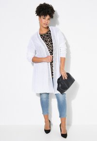 Vit öppet stickad cardigan över en svart mönstrad topp, i kombination med ljusblå jeans och svarta högklackade skor. Håller en svart kuvertväska.
