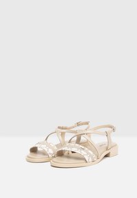 Sandali in pelle beige con cinturini incrociati, fibbia alla caviglia regolabile, tacco piatto e dettagli tessili decorati sulle fasce delle dita.