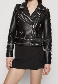 Veste de biker en cuir noir avec des clous en métal, fermeture éclair asymétrique et taille ceinturée. Associée à une mini-jupe noire avec des détails en dentelle.