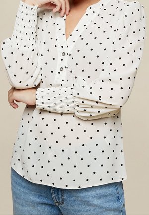 Žena nosící bílou halenku s dlouhým rukávem a černými puntíky s nařasenými manžetami, spárovanou s modrými džíny z denimu.