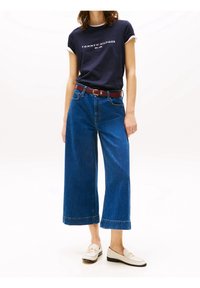 Granatowa koszulka z białym wykończeniem i logo, zestawiona z szerokimi, niebieskimi dżinsami oraz bordowym paskiem. Obuwie to jasne mokasyny.