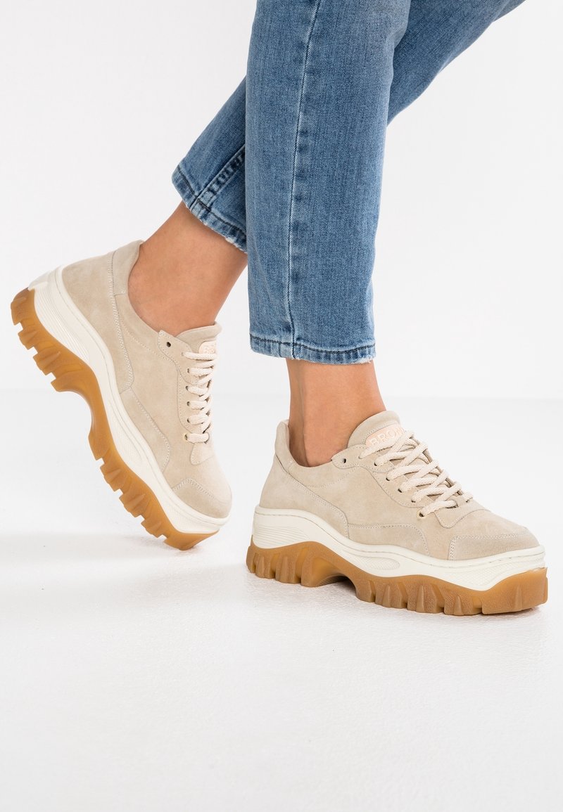 Beige Wildleder-Sneaker mit einer dicken weißen Mittelsohle und einer strukturierten Gummiaußensohle. Verfügt über runde Schnürsenkel und ein niedrig geschnittenes Design.