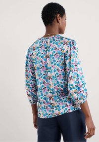 Blusa floreale con maniche a tre quarti, caratterizzata da un motivo nei toni pastello di blu, rosa e verde su un tessuto leggero, con un design del collo arricciato.