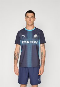 Camisa desportiva azul escura com detalhes em azul claro, apresentando um padrão em gradiente, logótipo da CMA CGM e marca Puma. Usada com calções azul-marinho.