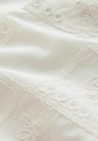 Tissu blanc avec une broderie délicate florale et géométrique dans un fil assorti, créant un motif texturé sur la surface.