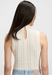 Młoda kobieta z długimi, ciemnymi włosami nosi bezrękawnik w kremowym kolorze z wzorem z koronką oraz z zapięciem na guziki z tyłu i jasnoniebieskie dżinsy.