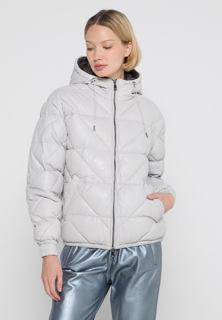 Mujer vestida con una chaqueta acolchada con capucha de color gris claro y cremallera, y pantalones brillantes de color azul, de pie contra un fondo blanco liso.