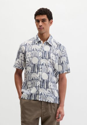 Mann trägt kurzärmeliges, zum Knöpfen geeignetes Hemd mit abstraktem Blumen- und geometrischem blau-weißem Muster, kombiniert mit brauner Hose, steht vor weißem Hintergrund.