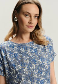 Kwiatowa bluzka w kolorze kremowym z niebieskim wzorem paisley, z krótkimi rękawami i okrągłym dekoltem, charakteryzująca się gładką teksturą i luźnym krojem.