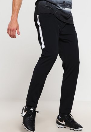 Homme portant un pantalon de sport noir avec une bande blanche sur le côté et des crampons noirs, debout sur un sol clair contre un mur uni.