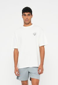 Λευκό βαμβακερό t-shirt με κοντά μανίκια, στρογγυλή λαιμόκοψη και μαύρο λογότυπο στο αριστερό στήθος. Συνδυάζεται με ανοιχτό γκρι σορτσάκι.