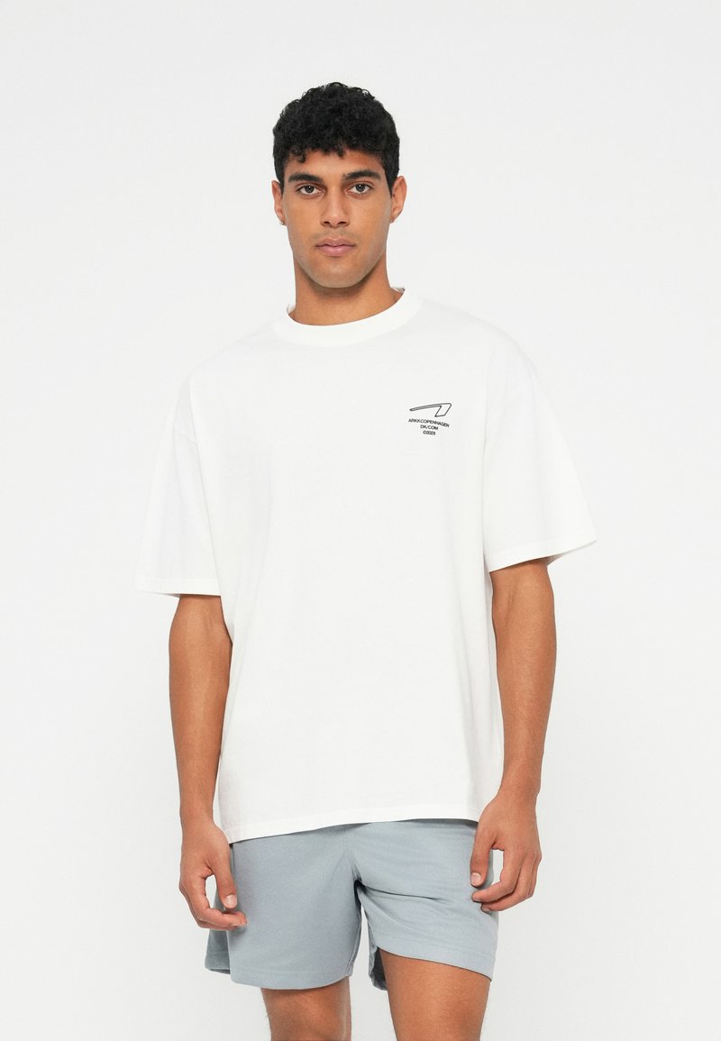 Λευκό βαμβακερό t-shirt με κοντά μανίκια, στρογγυλή λαιμόκοψη και μαύρο λογότυπο στο αριστερό στήθος. Συνδυάζεται με ανοιχτό γκρι σορτσάκι.