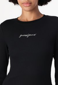 Juodas ilgas rankoves palaidinė, pagaminta iš raukšlėto audinio, su baltu siuvinėti "guessjeans" logotipu ant krūtinės. Apvali iškirptė ir prigludusi siluetas.