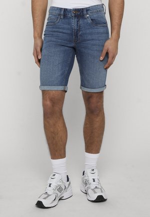 Homme portant un short en jean bleu retroussé, des chaussettes blanches et des chaussures de course blanches New Balance, debout devant un fond clair uni.