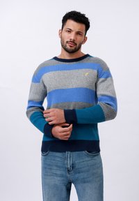 Jersey de punto a rayas en azul, gris y marino. Presenta una textura de canalé y un cuello redondo. Pequeño emblema en el pecho.
