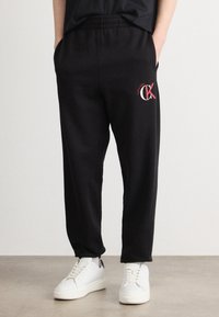 Μαύρες φόρμες με ελαστική μέση, πλαϊνές τσέπες και κόκκινο λογότυπο Calvin Klein στο μπούστο, σε συνδυασμό με λευκά αθλητικά παπούτσια.