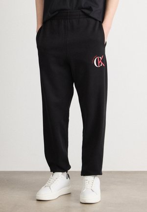 Czarne dresy z elastycznym pasem, kieszeniami bocznymi i czerwonym logo Calvin Klein na udzie, zestawione z białymi trampkami.