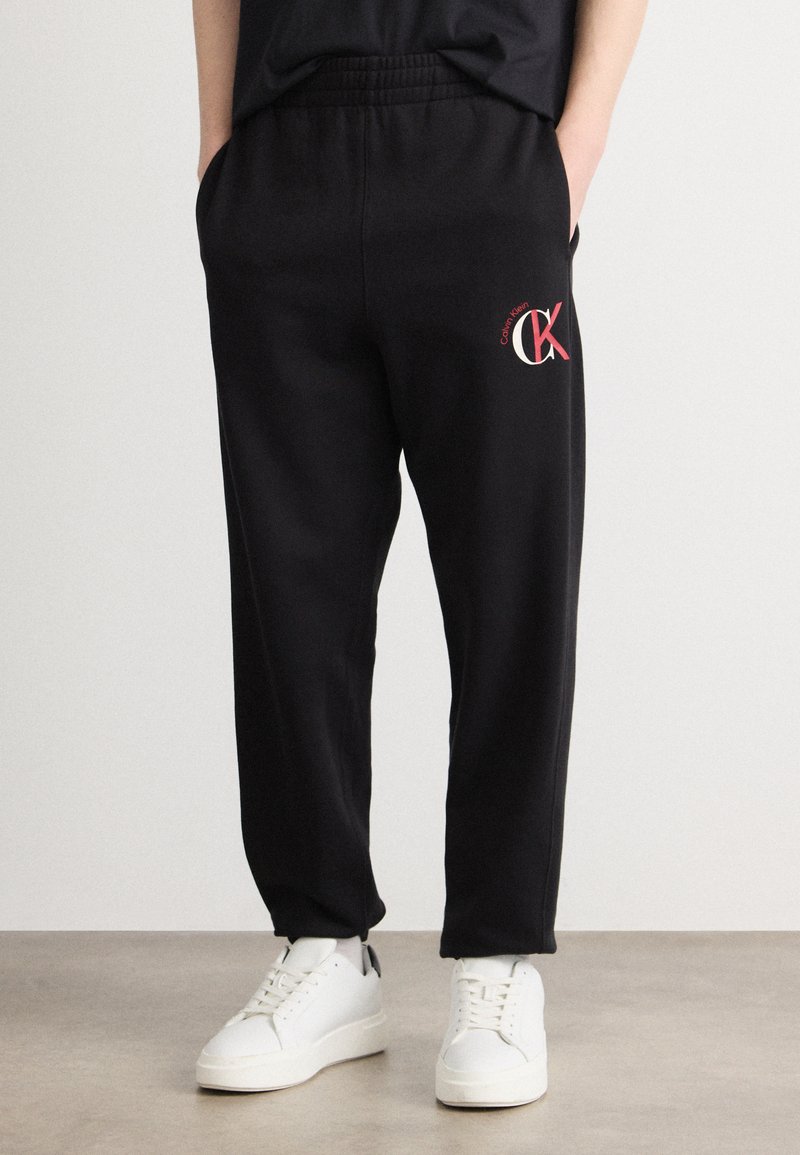 Μαύρες φόρμες με ελαστική μέση, πλαϊνές τσέπες και κόκκινο λογότυπο Calvin Klein στο μπούστο, σε συνδυασμό με λευκά αθλητικά παπούτσια.