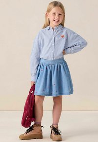 Chemise à rayures bleues et blanches avec un petit cœur rouge, associée à une jupe en denim ornée de motifs de cœurs rouges. Bottines en cuir marron à lacets et sac rouge.