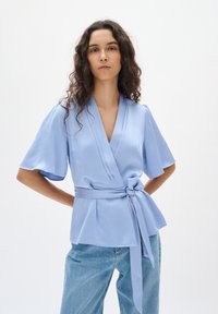 Blusa avvolgente di colore blu chiaro con maniche corte e svasate. Presenta una cintura annodata in vita, una texture strutturata e un design a scollo a V. Abbinata a denim.