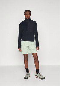 Giacca in pile blu navy con cerniera abbinata a pantaloncini cargo verde menta, arricchiti da tasche. Scarpe sportive grigie coordinate e calzini neri.
