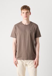 Braunes Baumwoll-T-Shirt mit Rundhalsausschnitt und kurzen Ärmeln, versehen mit einem kleinen schwarzen Logo auf der Brust. Sanfte Textur, lockere Passform.