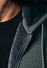 Grå zip-up hoodie med plysfor, struktureret stof og en tætsiddende sort indershirt. Har en glat lynlås og kontrastsyet.
