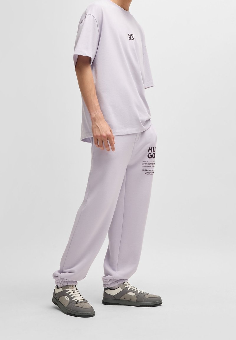 Person wearing light purple oversized T-shirt and matching joggers with branded text, paired with gray and white sneakers on a plain background.
