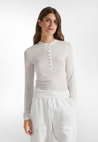 Maglietta bianca a maniche lunghe realizzata in tessuto leggero, con scollatura rotonda e bottoni. Abbinata a pantaloni bianchi larghi per un look rilassato.