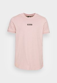Lyserød bomulds T-shirt med rund hals, korte ærmer, og et sort logo-patch på brystet. Glat tekstur og almindelig pasform.