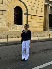 Marinblå kofta med knappar, vita vida byxor och bruna loafers; står på en texturerad asfaltyta nära en stenbyggnad.