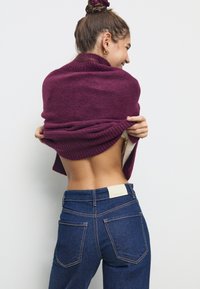 Bordo pleten pulover premeten čez ramena, v kombinaciji s temno modrimi kavbojkami. Ima rebraste manšete in oznako na pasu.