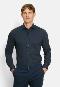 Navyblauwe lange mouwen shirt met witte stippen, met een knoopsluiting kraag en een slanke pasvorm. Gepaard met donkere broek, handen in de zakken.
