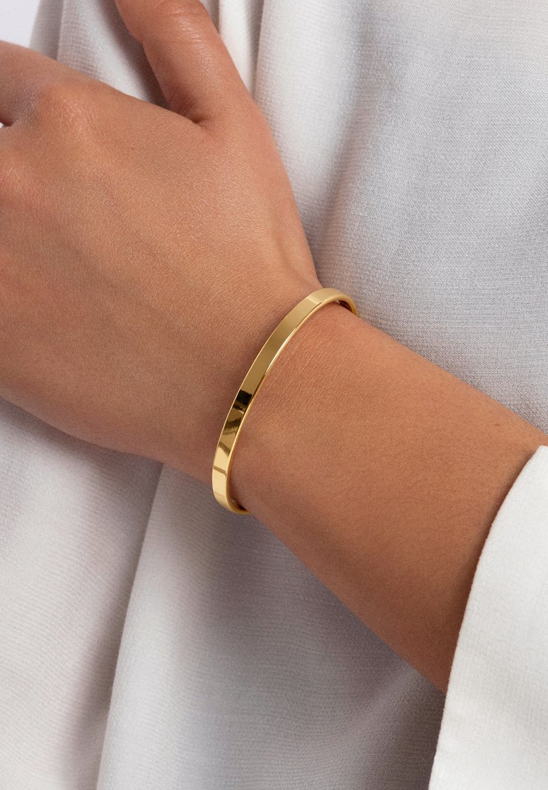 Main gauche portant un bracelet en or lisse et brillant au poignet sur un fond de tissu blanc.