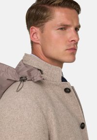 Beidzieles vilnas mētelis ar augstu apkakli, kas papildināts ar pelēku kapuci, melniem pogām un strukturētu apdari. Sānu detaļās redzama aparatūra un aukla.