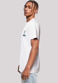 White cotton t-shirt featuring a round neck, short sleeves, and a printed logo on the left chest area. Styled with light blue denim jeans.
